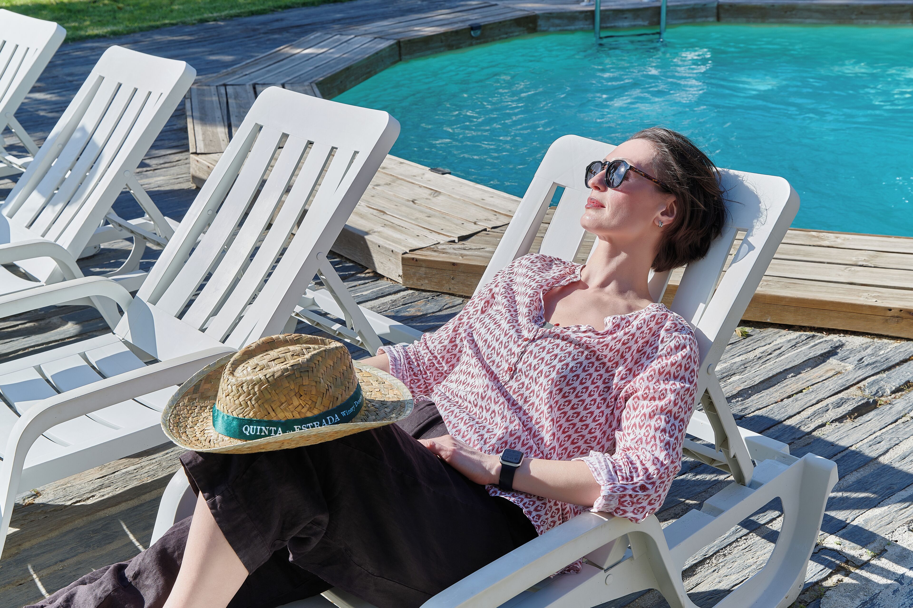 A person relaxing on a white lounge chair by a pool with a straw hat and a watch.