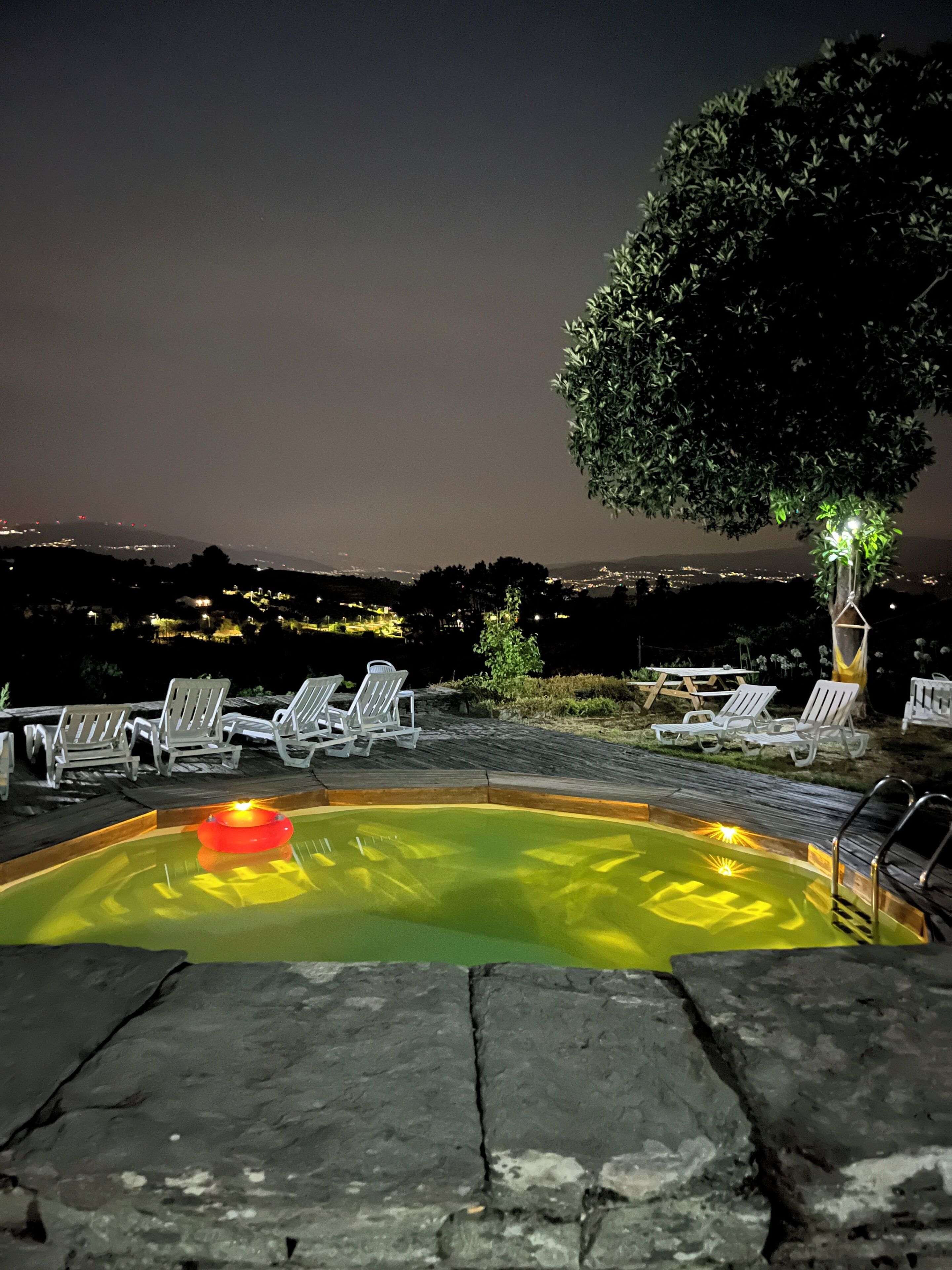 Una piscina con sillas de descanso, un árbol y vista a un paisaje urbano distante en la noche.