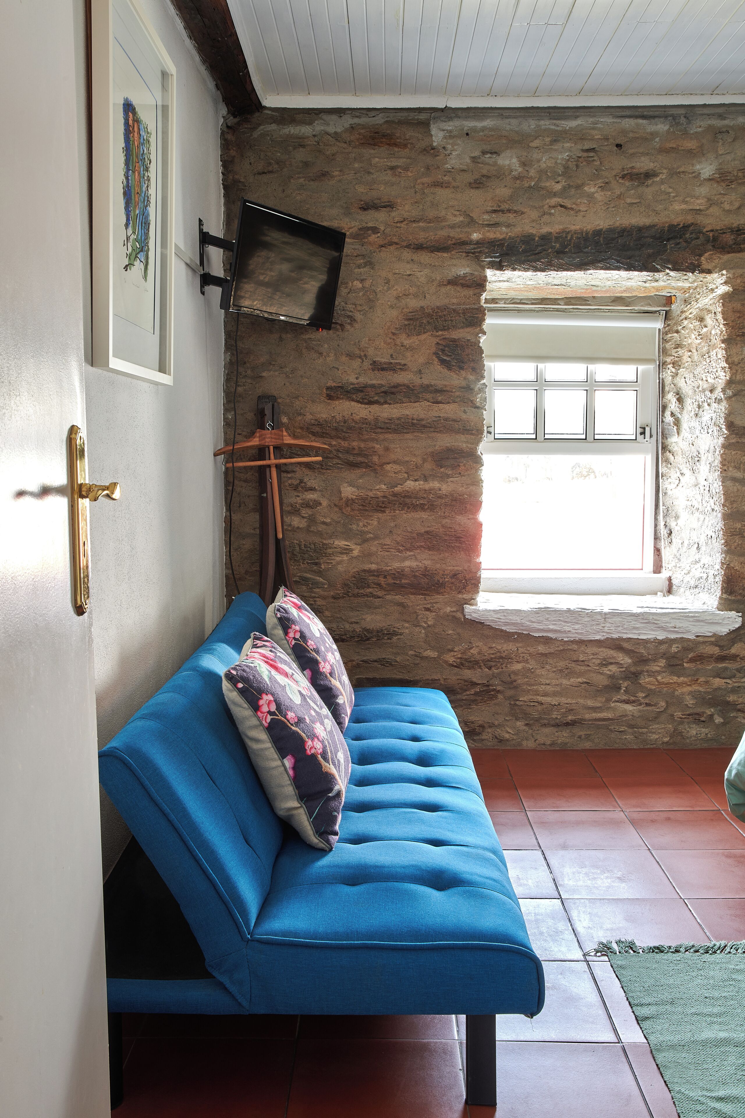 A room with a blue sofa, a wooden door, a stone wall, a window, and a television mounted on the wall.