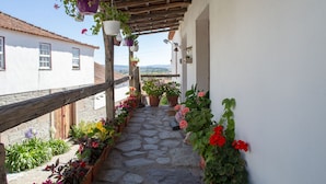 A stone-paved balcony with hanging flower pots and potted plants, overlooking a rural landscape.