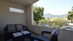 A balcony with a sofa, a small table, and a chair. The balcony overlooks a mountainous landscape with trees and a clear sky. - Cozy apartment in Moriani Plage (Moriani Plage)