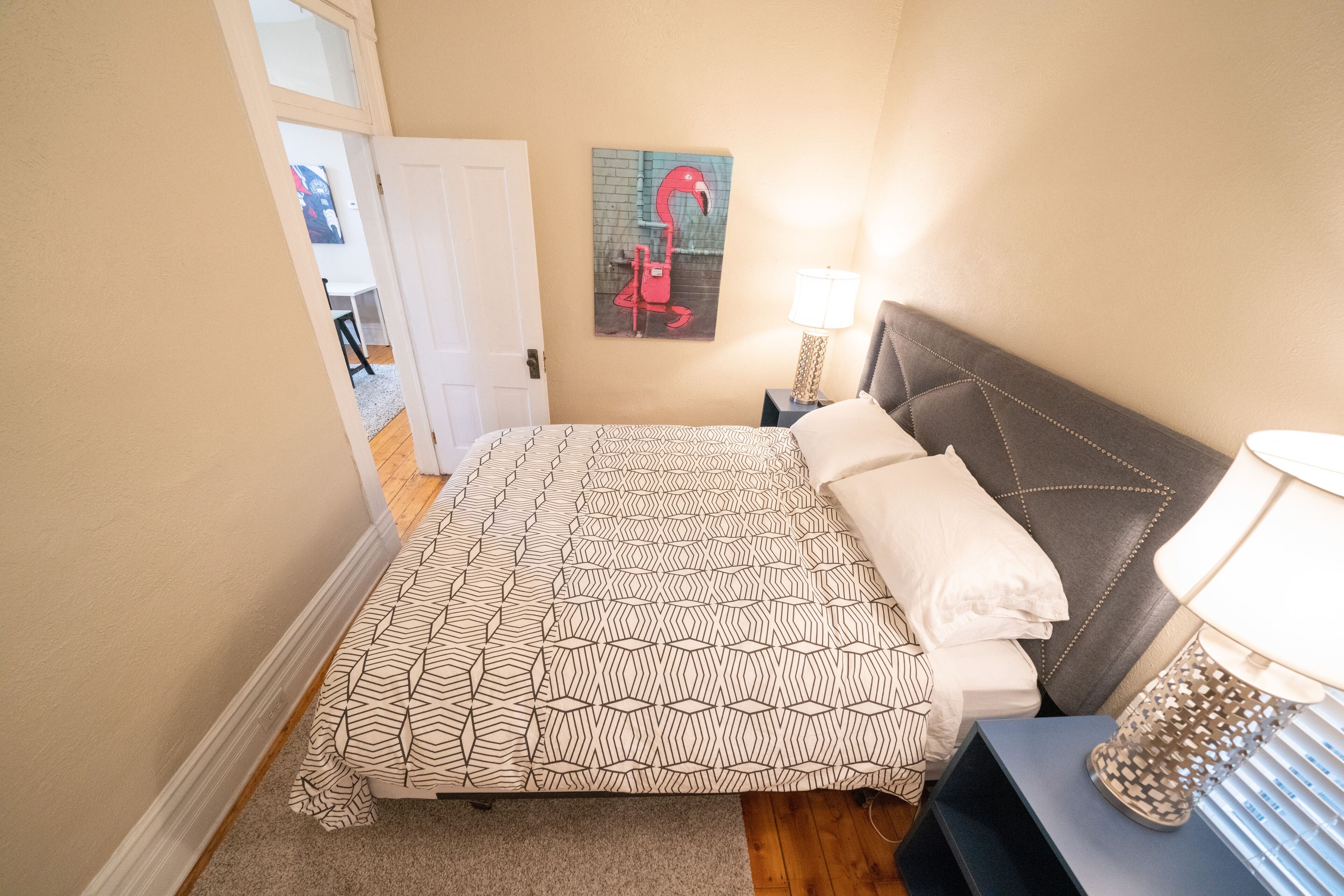A bedroom with a patterned bedspread, a nightstand with a lamp, and a painting of a flamingo on the wall.