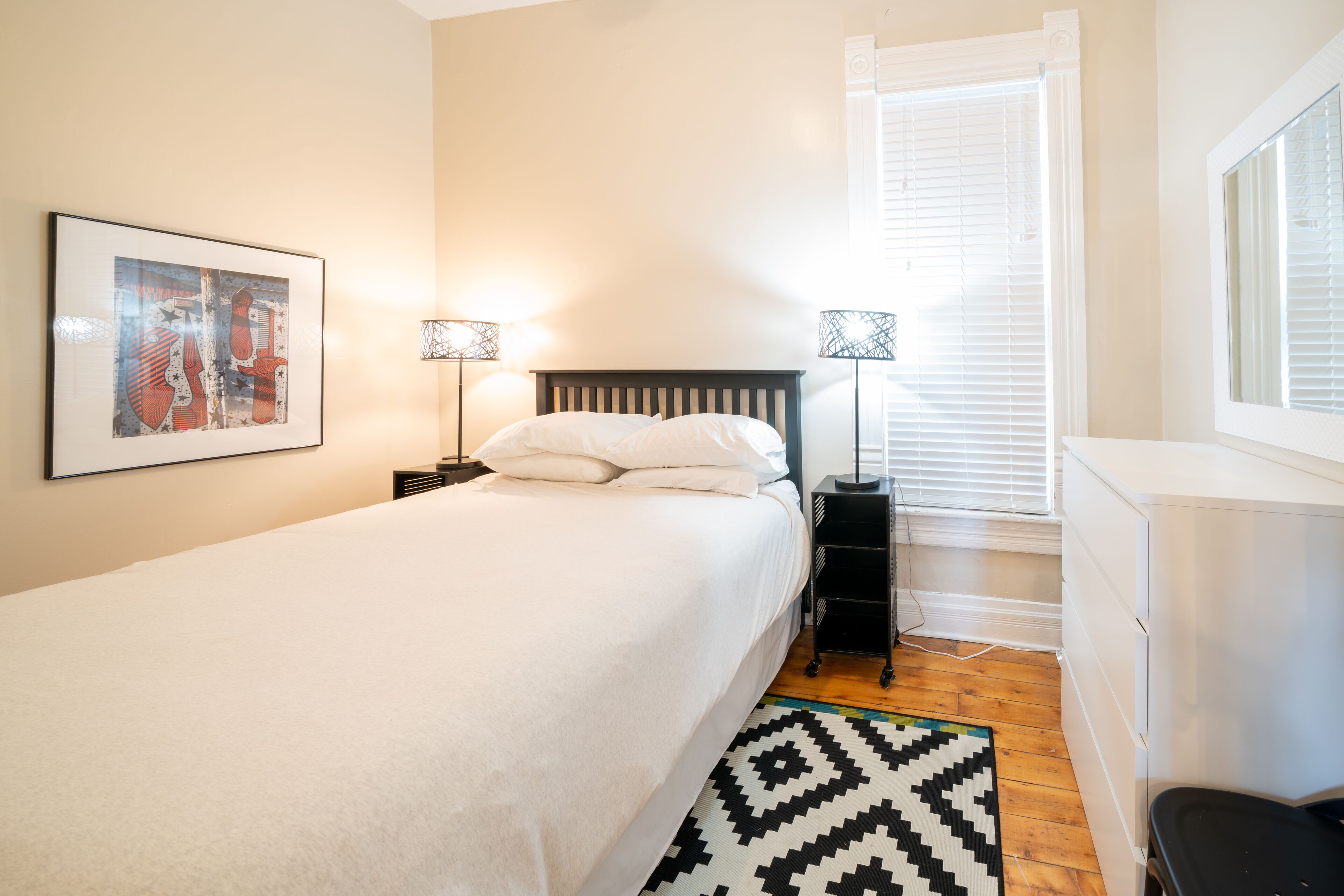A bedroom with a bed, two bedside tables with lamps, a window with blinds, a radiator, and a wall-mounted picture.