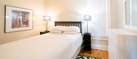 A bedroom with a bed, two bedside tables with lamps, a window with blinds, a radiator, and a wall-mounted picture.