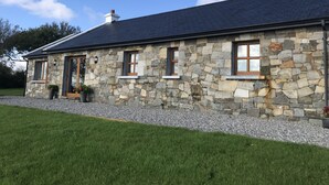 A stone house with a dark roof, multiple windows, and a gravel driveway. - Mullach Cottage (Tourmakeady)