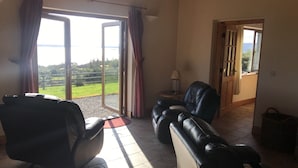 A room with a large sliding glass door leading to a balcony, a leather sofa, a small table with a lamp, and a wooden door leading to another room. - Mullach Cottage (Tourmakeady)