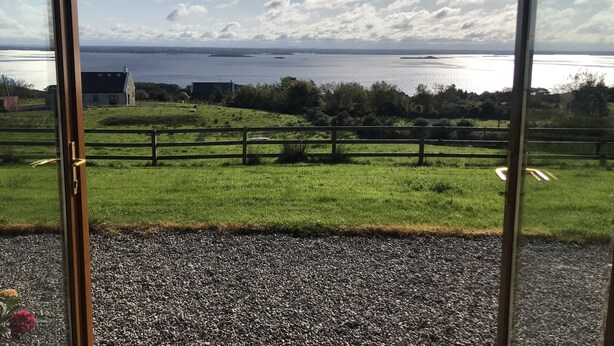 A view from a room with a large window overlooking a grassy area with a fence, a body of water, and distant landforms. - Mullach Cottage (Tourmakeady)