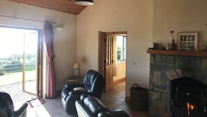 A cozy living room with a fireplace, leather sofas, a stone fireplace surround, and a view through a glass door. - Mullach Cottage (Tourmakeady)