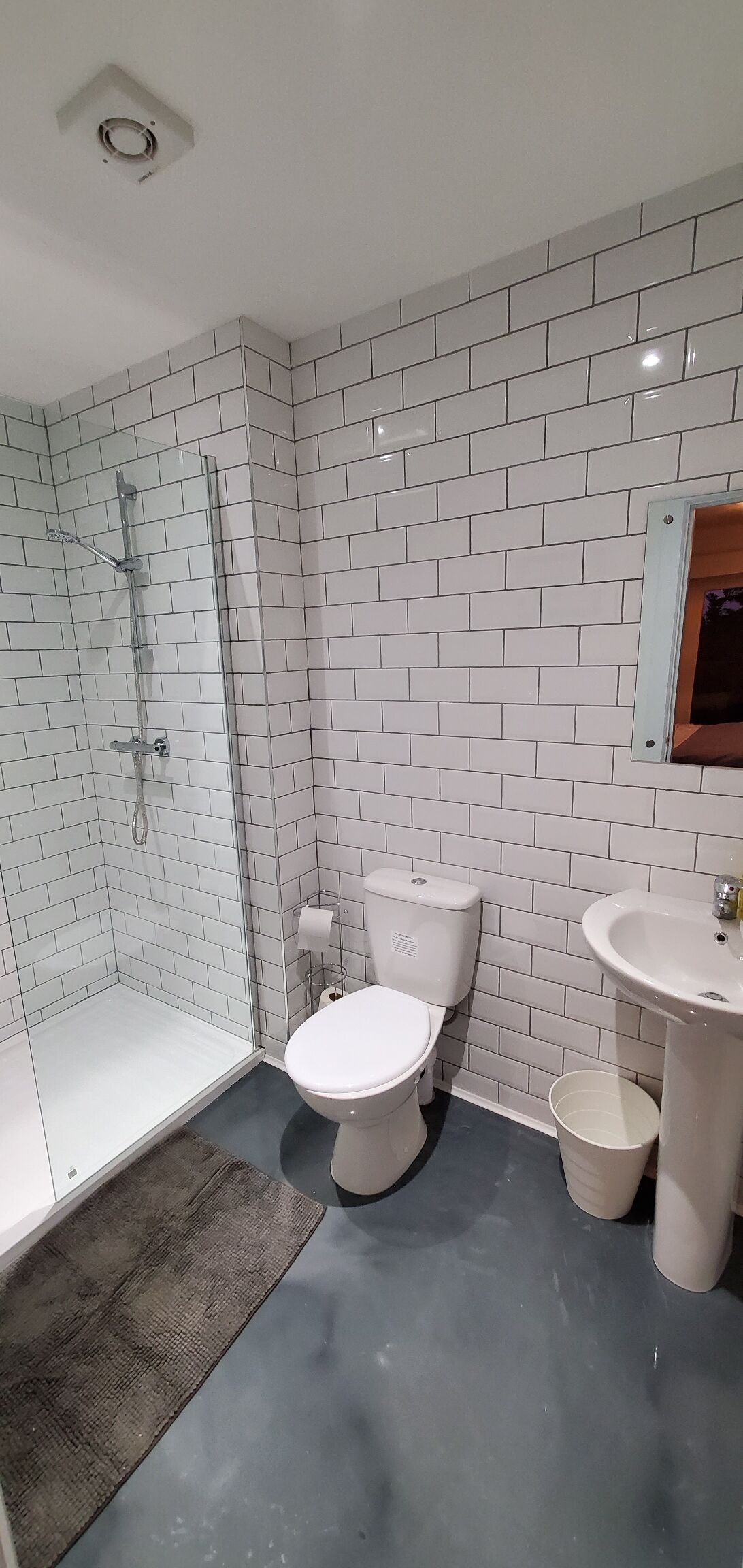 Un baño con paredes de azulejos blancos, cabina de ducha de vidrio, inodoro, lavabo y un espejo pequeño.