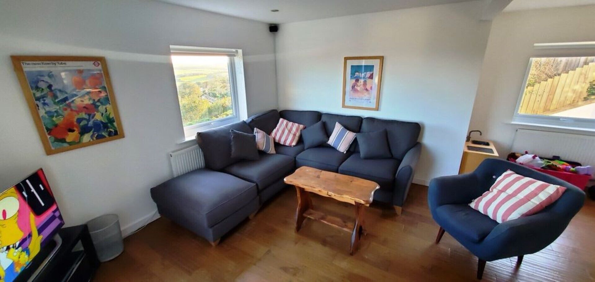 A modern living room with a sectional sofa, a wooden coffee table, and a blue armchair. There are two windows providing natural light, and artwork is displayed on the walls.