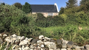 Een stenen huis met een donker dak, omringd door weelderige begroeiing en een stenen muur op de voorgrond.