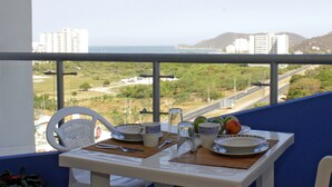 A balcony with a table set for two, overlooking a coastal landscape with buildings and a road. - Apartamento de Yuly Cerca a la Playa Sector Bello Horizonte Santa Marta  (Santa Marta)