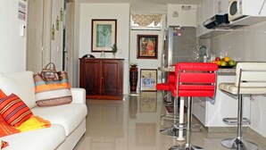 A modern kitchen with a refrigerator, microwave, and fruit bowl. There is a red bar stool and a white bar counter. A wooden cabinet and a framed picture are visible in the background. - Apartamento de Yuly Cerca a la Playa Sector Bello Horizonte Santa Marta  (Santa Marta)