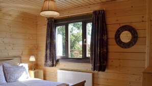 A wooden cabin bedroom with a bed, a nightstand, a lamp, a window with curtains, and a radiator. - 3 bedroom accommodation in Nomansland, near Tiverton (Nomansland, near Tiverton)