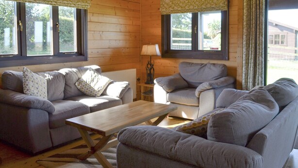 A cozy living room with a sofa set, a coffee table, and a lamp. There are windows with curtains and a view of the outside. - 3 bedroom accommodation in Nomansland, near Tiverton (Nomansland, near Tiverton)