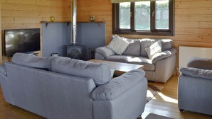 A log cabin interior with a fireplace, two sofas, a television, and a window with a curtain. - 3 bedroom accommodation in Nomansland, near Tiverton (Nomansland, near Tiverton)