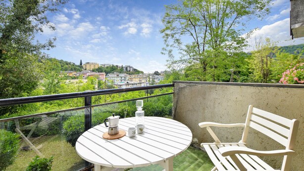 Un balcón con una mesa redonda y una silla blanca, con vistas a un paisaje urbano con edificios y áreas verdes.