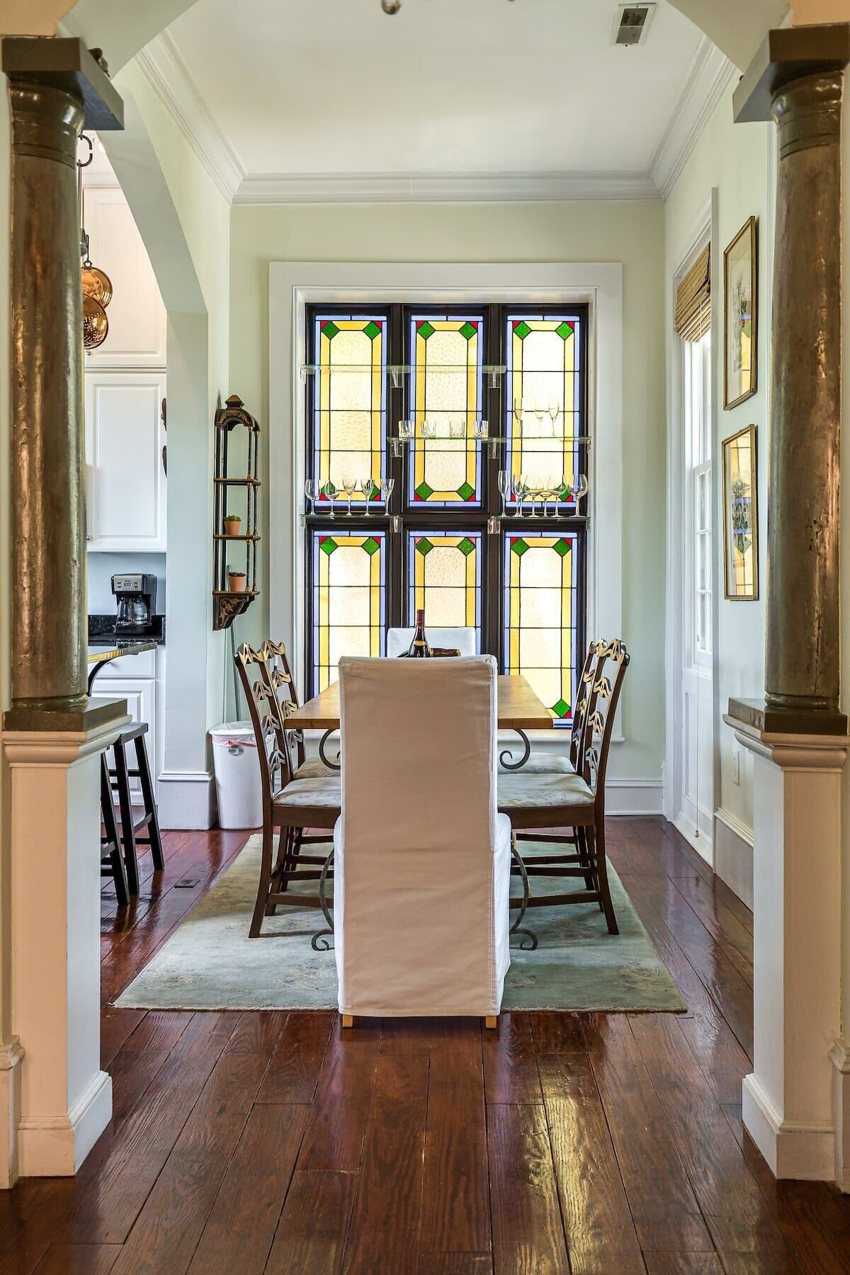 A dining area with a table and chairs, a window with stained glass, and a kitchen in the background.