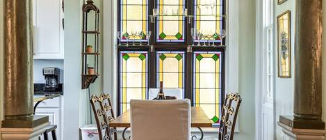 A dining area with a table and chairs, a window with stained glass, and a kitchen in the background.