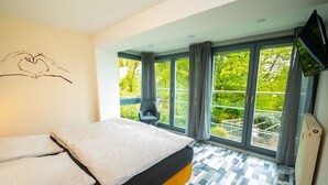 A modern hotel room with a large bed, a chair, a TV, and a balcony with a view of greenery.