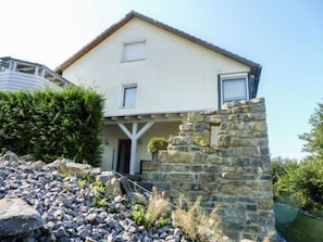 Una casa de dos pisos con fachada de piedra, un pequeño porche y un jardín con rocas y arbustos.