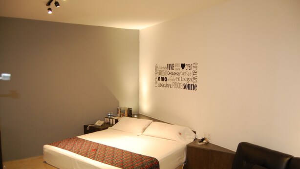 Una cama bien hecha con un edredón estampado, una mesita de noche con una lámpara y una pared con mensajes motivacionales.