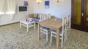Una habitación con una mesa de madera, sillas blancas, un sofá, un televisor montado en la pared y una puerta con un panel de vidrio decorado.