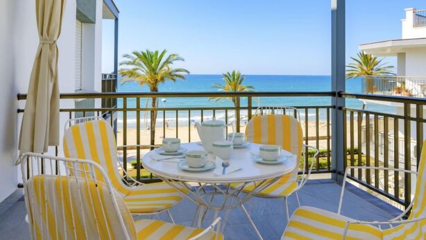 Un balcón con sillas a rayas amarillas y una mesa dispuesta para el té, con vistas a una playa con palmeras y el mar.