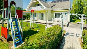 Ein zweistöckiges Haus mit einer Terrasse, einem Spielbereich mit Rutschen und Klettergerüsten sowie Solaranlagen auf dem Dach.