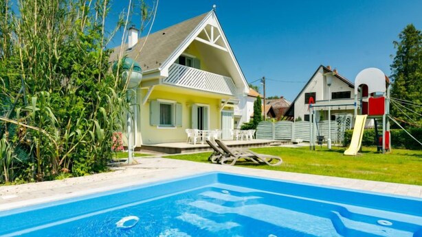 Ein Schwimmbecken mit klarem, blauem Wasser, umgeben von einer grünen Rasenfläche und einem Haus mit geneigtem Dach und weißer Fassade.