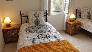 Ein Schlafzimmer mit zwei Betten, jeweils mit gemustertem Überwurf und passenden Kissen. An den Betten stehen Nachttische mit Lampen, ein Fenster bietet Blick auf Bäume und ein an der Wand befestigtes Schild ist zu sehen.