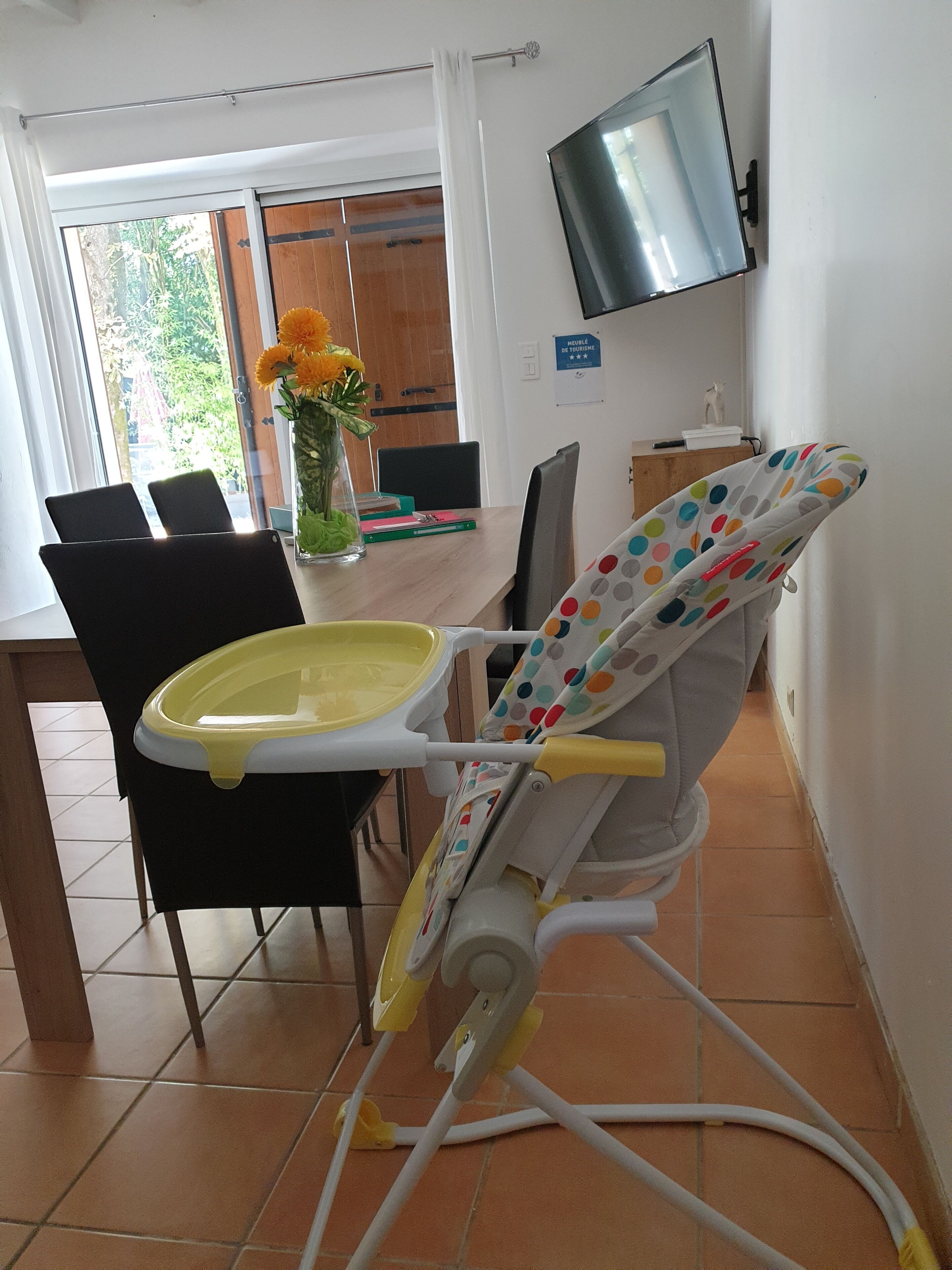 Ein Essbereich mit Hochstuhl, Stühlen, einem Tisch mit Büchern, einer Vase mit Blumen und einem Fernseher an der Wand.