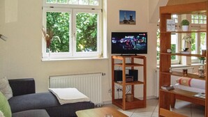 Un salon comprenant un canapé, une table basse, un téléviseur posé sur un meuble en bois, une fenêtre donnant sur un paysage verdoyant et une étagère ornée d’objets décoratifs.