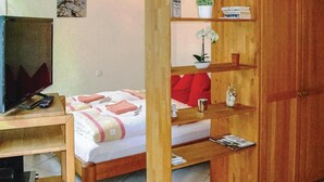 Une chambre avec un lit, un bureau en bois sur lequel se trouvent une télévision et une télécommande, une bibliothèque remplie de livres et d’objets décoratifs, et une petite plante en pot.