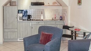 Un espace de vie compact comprenant un coin cuisine, un fauteuil gris agrémenté d’un coussin rouge, une petite table en bois et un coin repas avec une table en verre et des chaises noires.