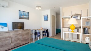 Una habitación de hotel con cama, escritorio con silla, televisión, lámpara y kitchenette.