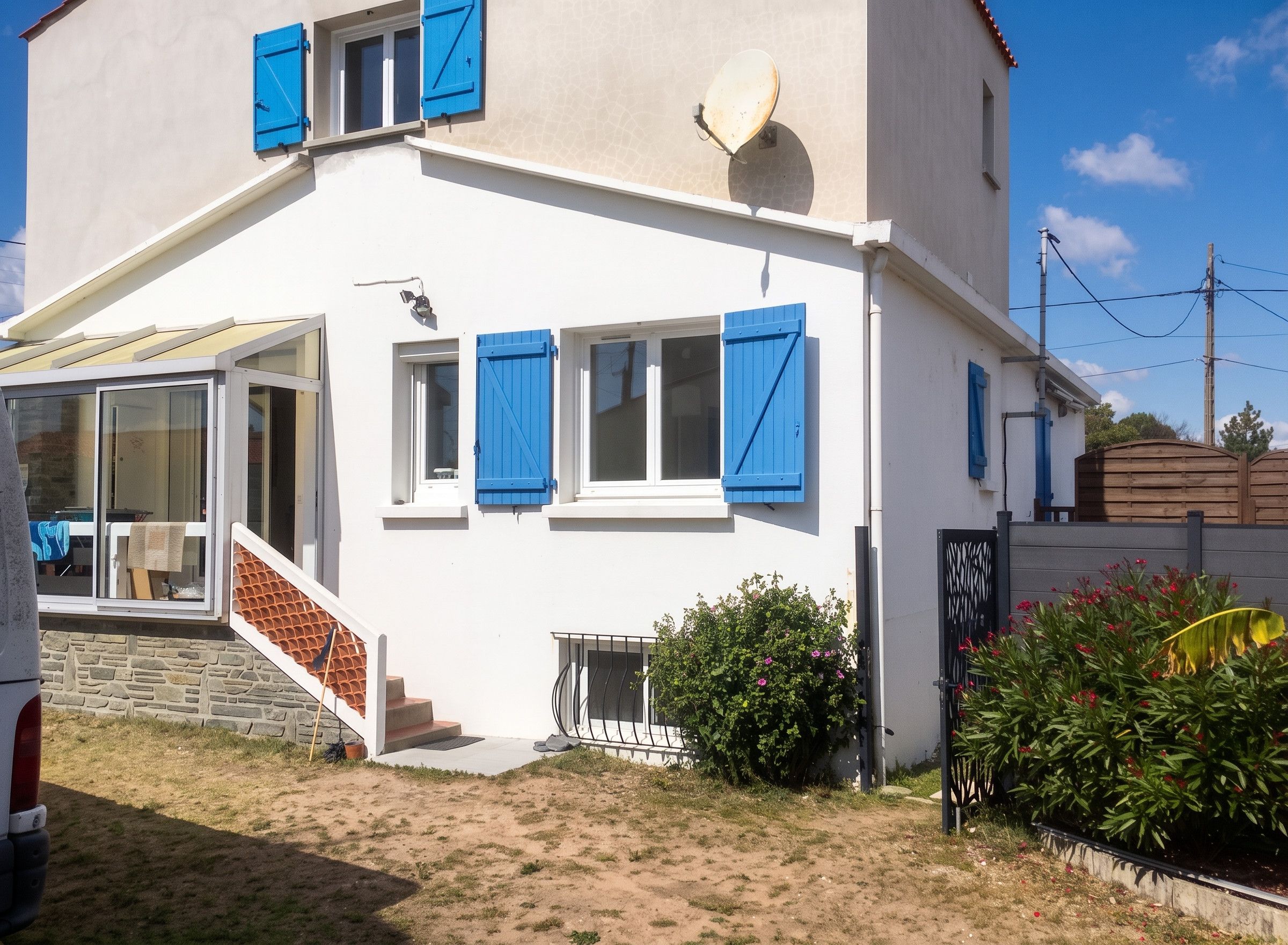 A white building with blue shutters, a satellite dish, and a small garden with red flowers.