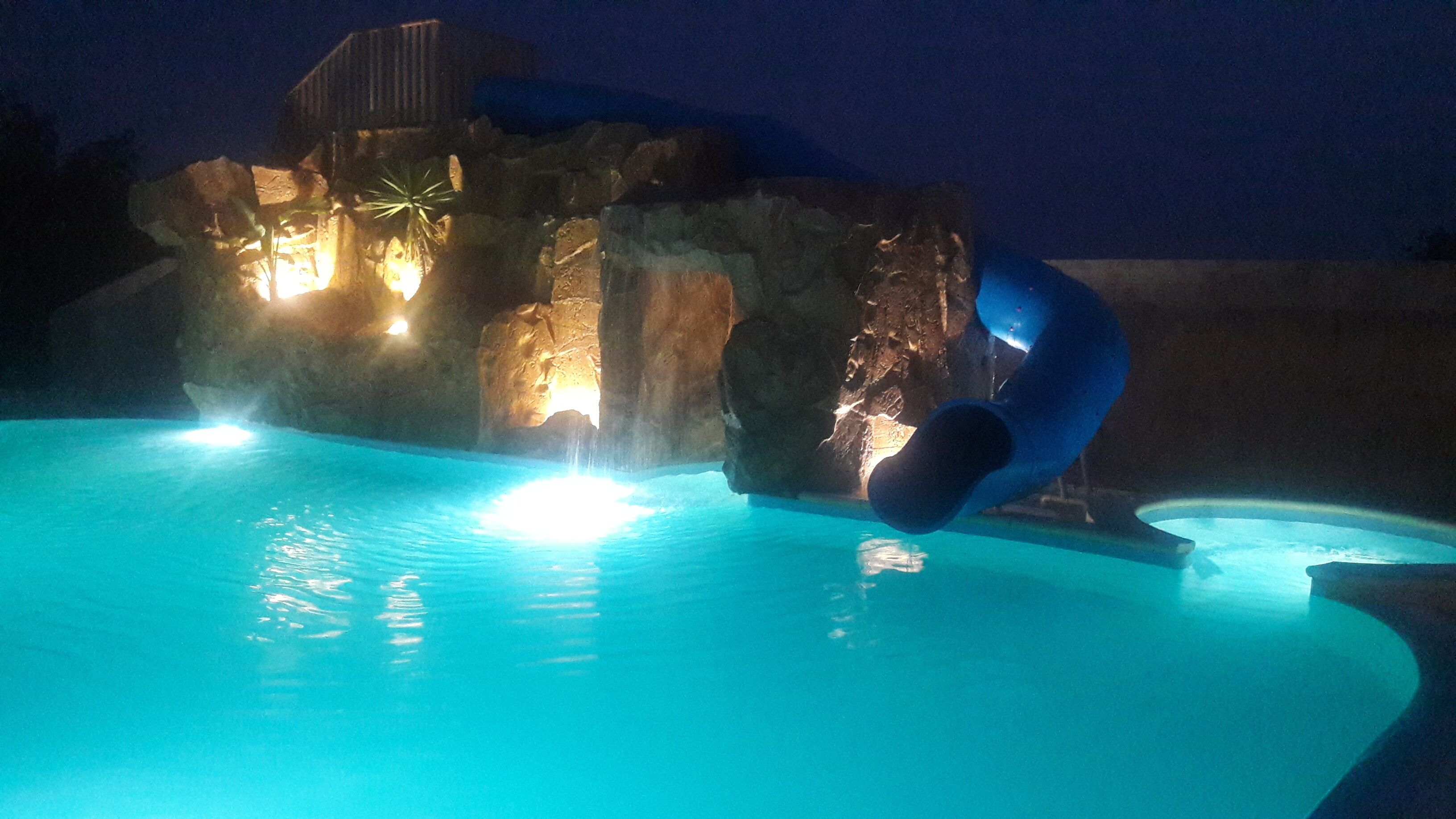 Una piscina con tobogán y formaciones rocosas iluminadas de noche.