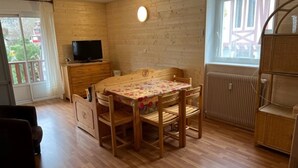 A cozy room with a wooden table and chairs, a television on a wooden cabinet, and a window with a view of a building outside. - Bright 2-Room with Balcony and WIFI - Village Center (Brides-les-Bains)