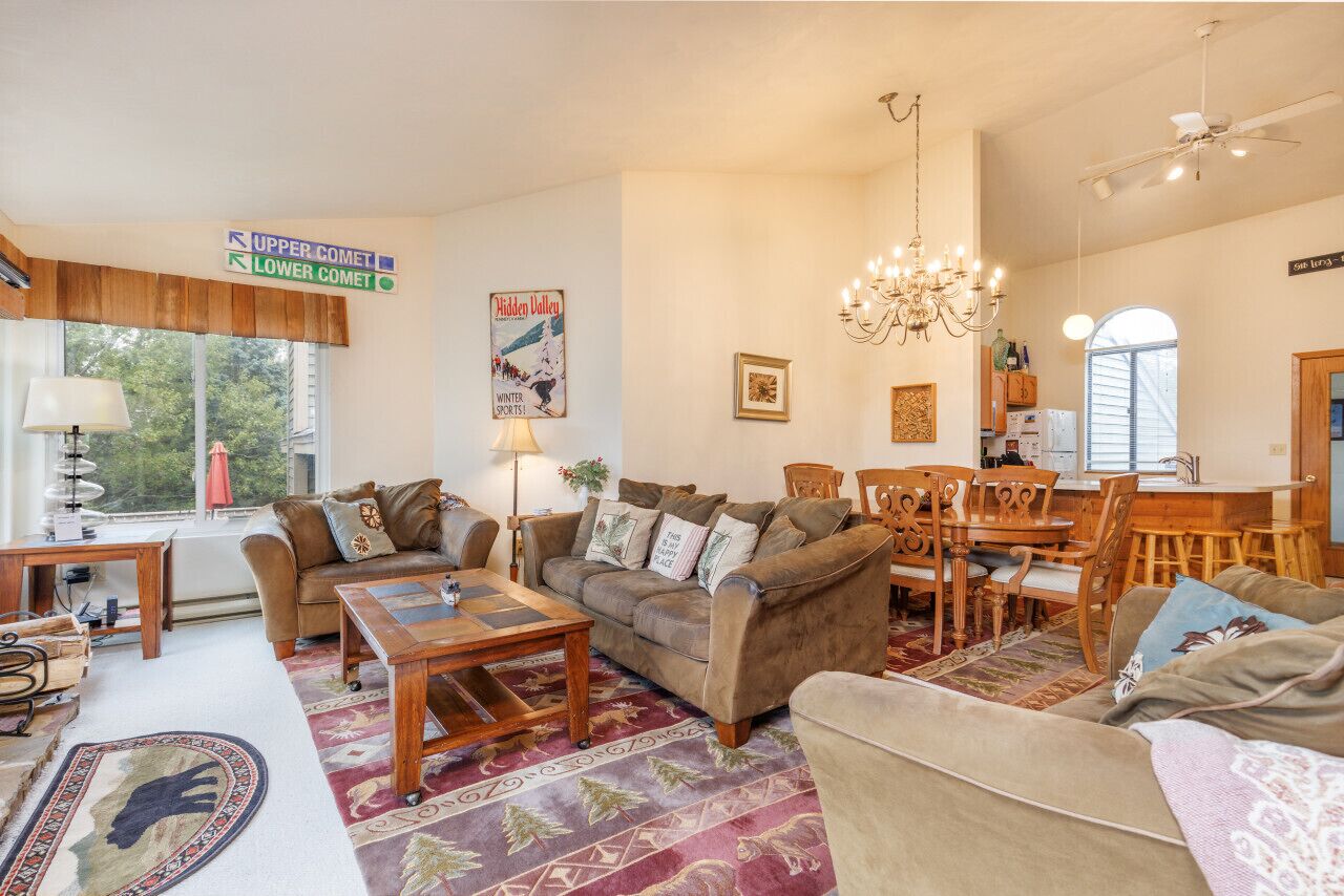 Een gezellige woonkamer met een bruine bank, een houten salontafel en een kroonluchter. Er is een eethoek met houten stoelen en een tafel.