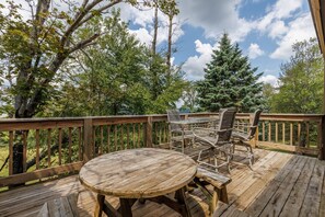 Une terrasse en bois avec une table ronde et des chaises, entourée d’arbres et sous un ciel dégagé.