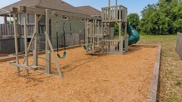 A playground with a slide and climbing structures, surrounded by a wooden fence and a house in the background.