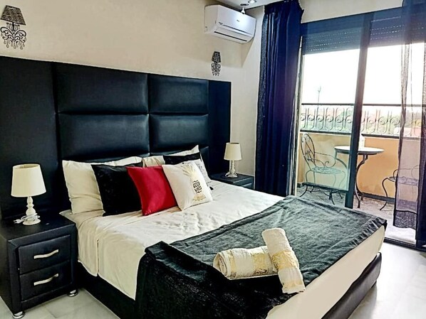 A hotel room with a large bed, two bedside tables with lamps, a balcony with a chair, and an air conditioning unit.