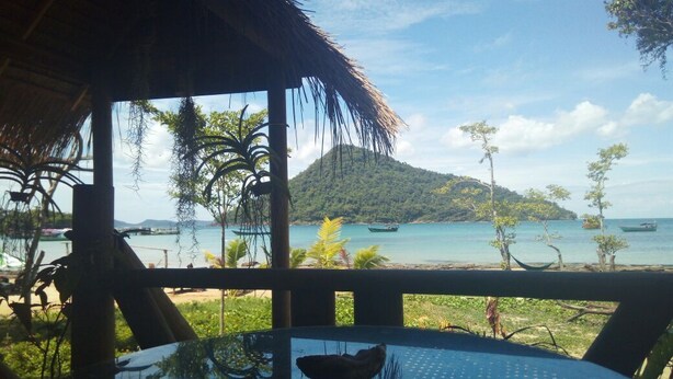 Vista de praia tropical de uma área coberta, com mesa e cadeiras, com vista para o mar e uma pequena ilha.