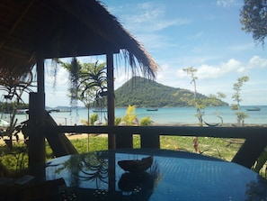 A tropical beach view from a covered area with a table and chairs, overlooking the sea and a small island. - Cupido Beach SPA Resort (Koh Rong Sanloem)