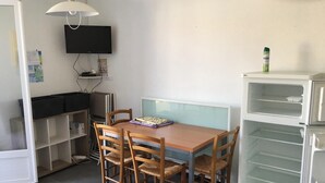 A compact room with a dining table, four wooden chairs, a flat-screen TV mounted on the wall, a white refrigerator, and a white cabinet with open shelves. - Homerez - House/Garden, Saint-Hilaire-de-Riez (Saint-Hilaire-de-Riez)