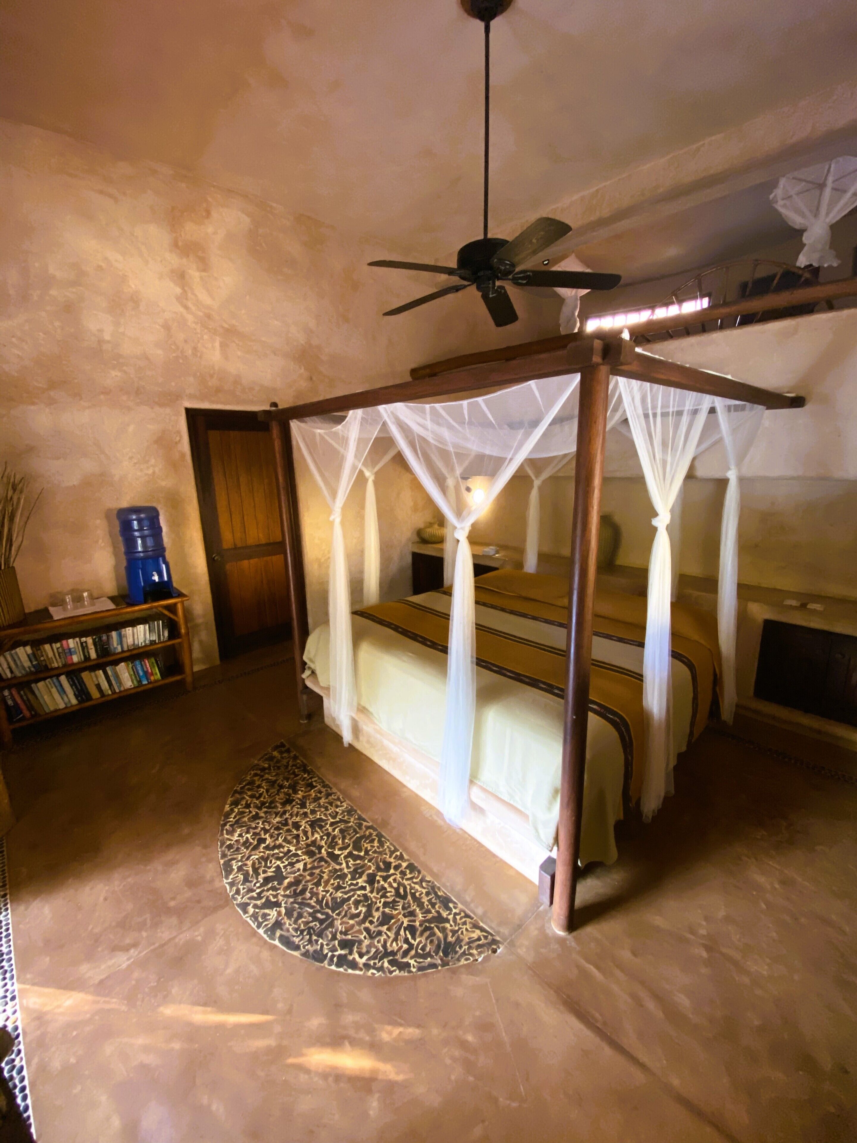 Una habitación con cama con dosel, ventilador de techo, estantería y alfombra estampada.