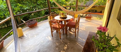A covered outdoor seating area with a table and chairs, surrounded by potted plants and a hammock.