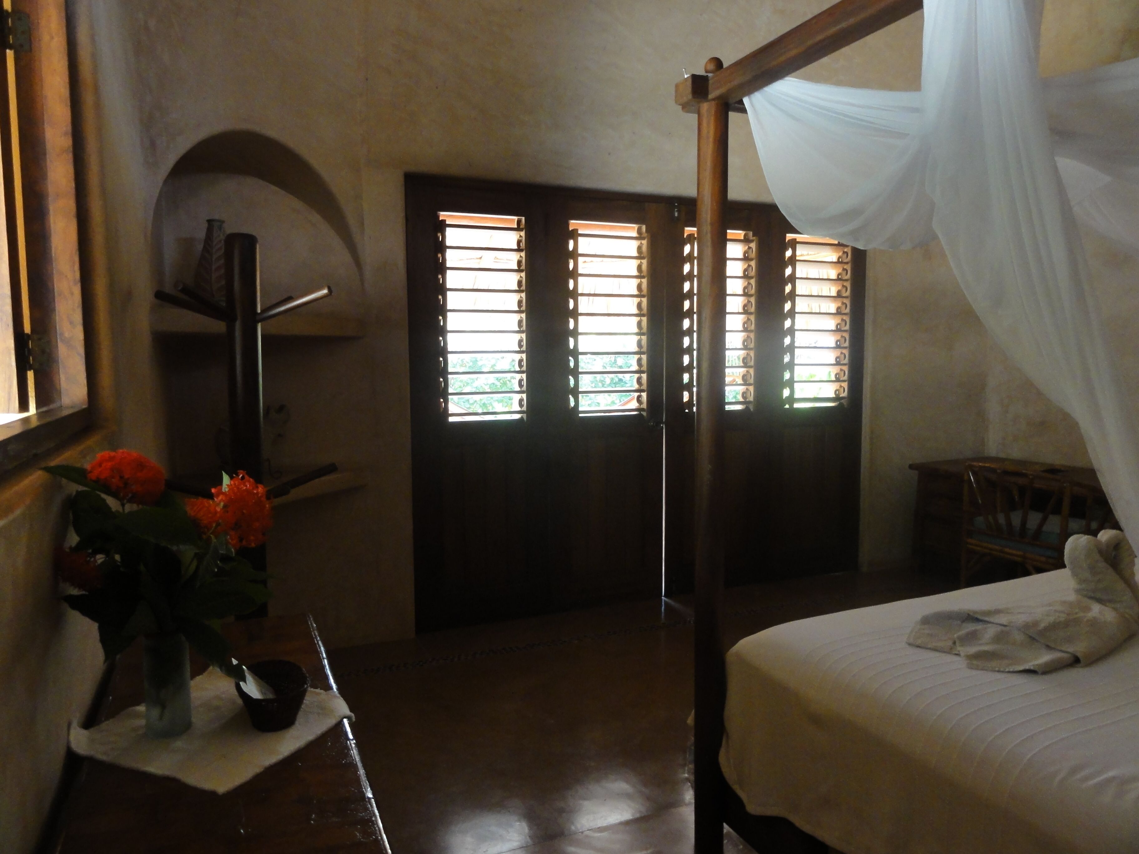 A bedroom with a four-poster bed, a nightstand with a vase of flowers, a wooden chair, and a window with blinds.