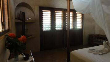 A bedroom with a four-poster bed, a nightstand with a vase of flowers, a wooden chair, and a window with blinds.